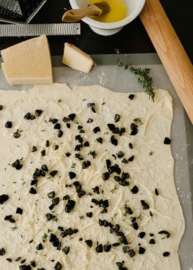 Sprinkling a sheet of rolled out puff pastry with chopped olives and grated cheese.