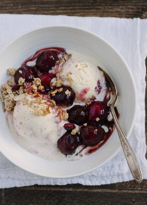 A white bowl filled with two scoops of vanilla icecream, with Cherry Wine Sauce drizzled on top, and granola sprinkled throughout.