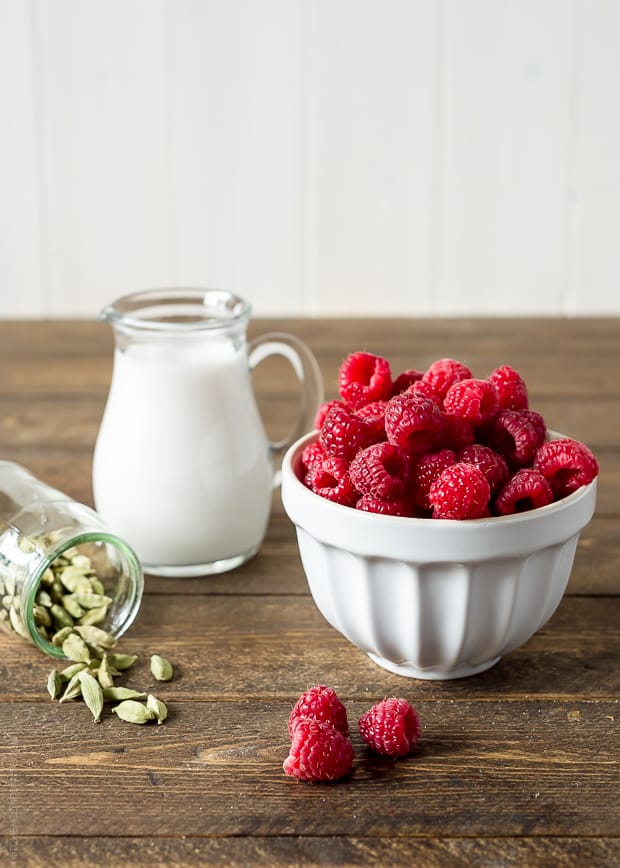 A small glass pitcher full of coconut cream, a white bowl full of fresh raspberries, and a jar of cardamom seeds spilling onto the countertop.