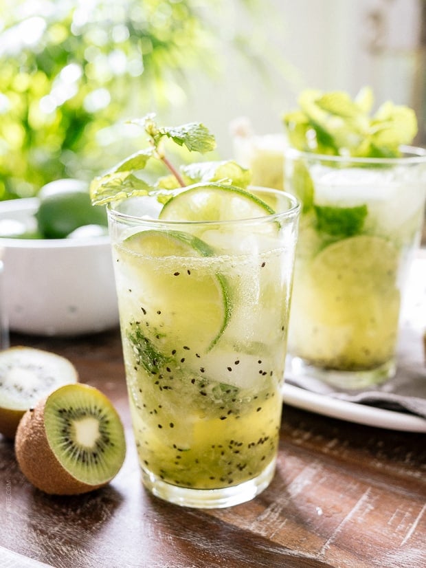 Kiwi Lime Mojitos on a wooden surface, surrounded by fresh kiwi and limes.