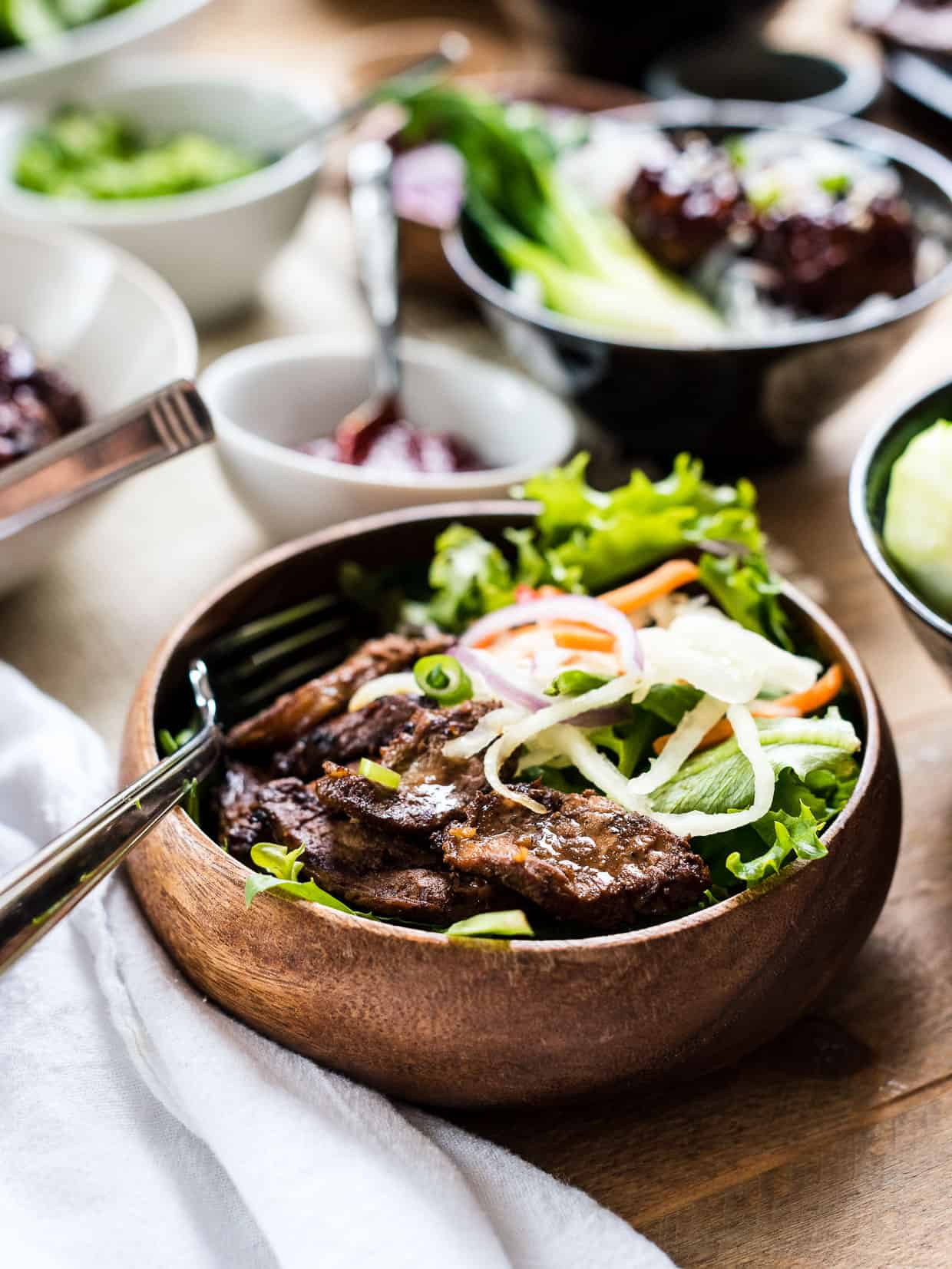 A wooden bowl filled with Filipino Pork BBQ, fresh greens, and lots of toppings.