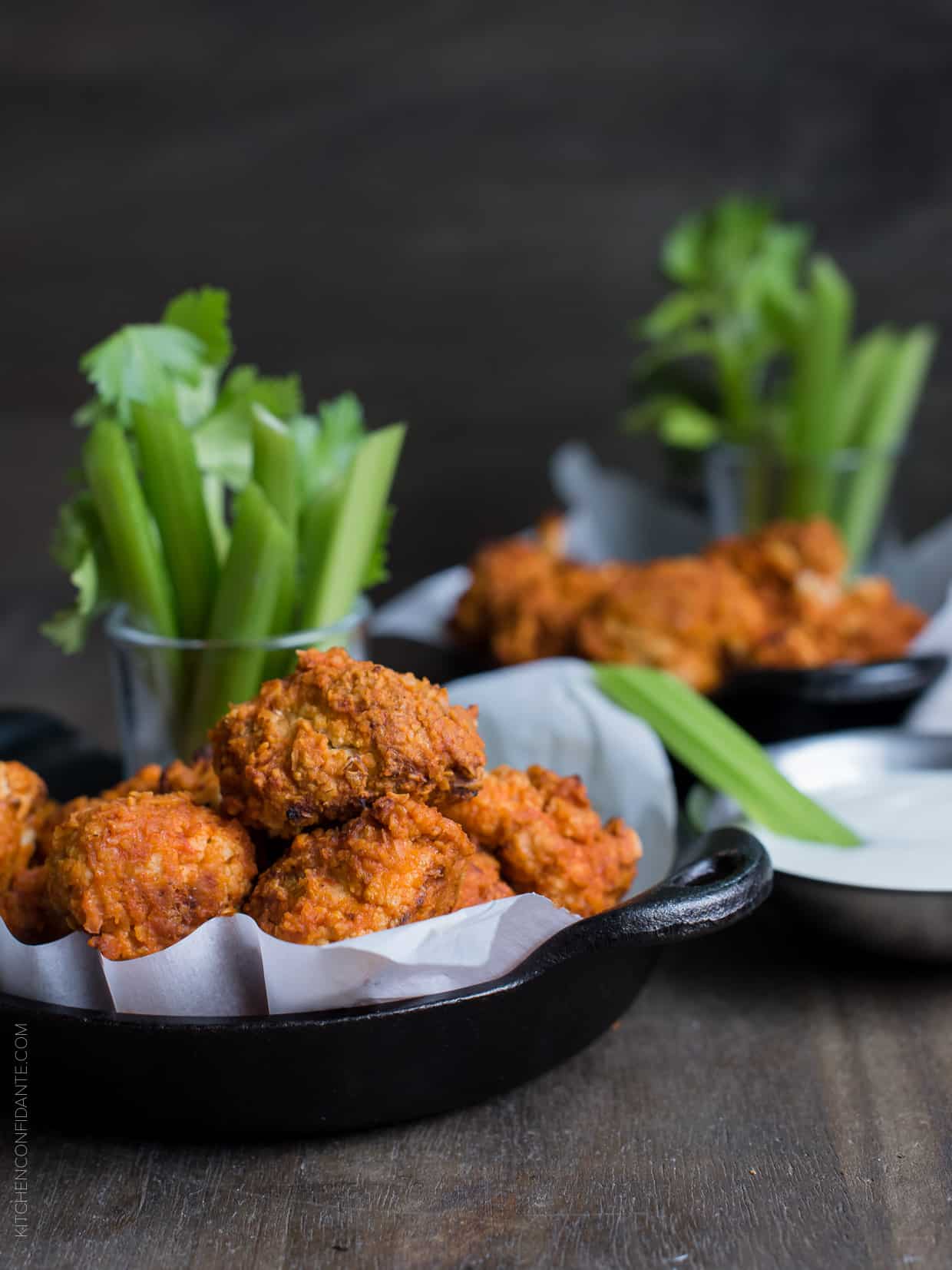 Hummus Battered Buffalo Cauliflower "Wings" served in small cast iron skillets alongside stalks of celery.