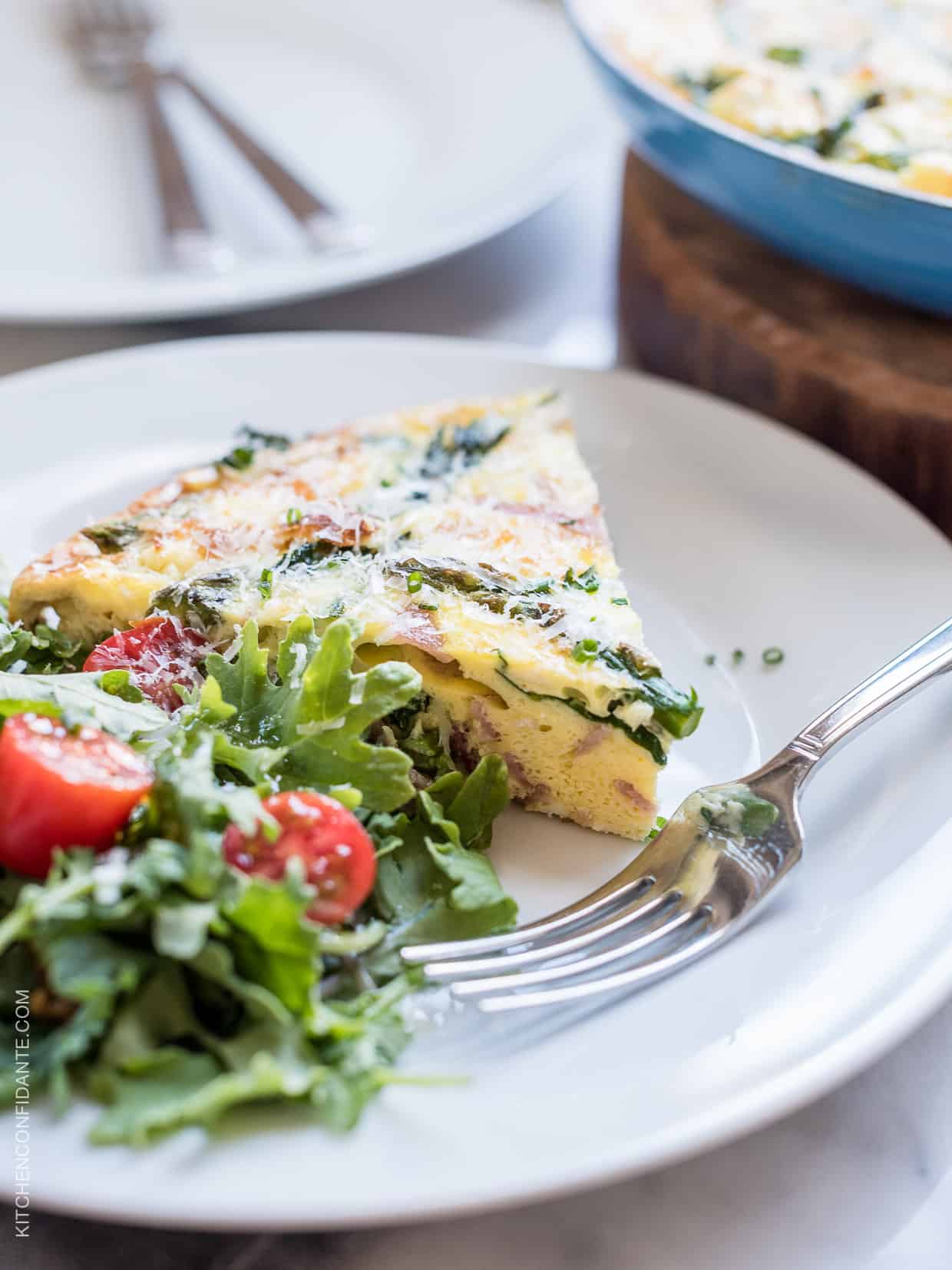 A wedge of Asparagus, Ham and Kale Frittata served on a white plate alongside a green salad.