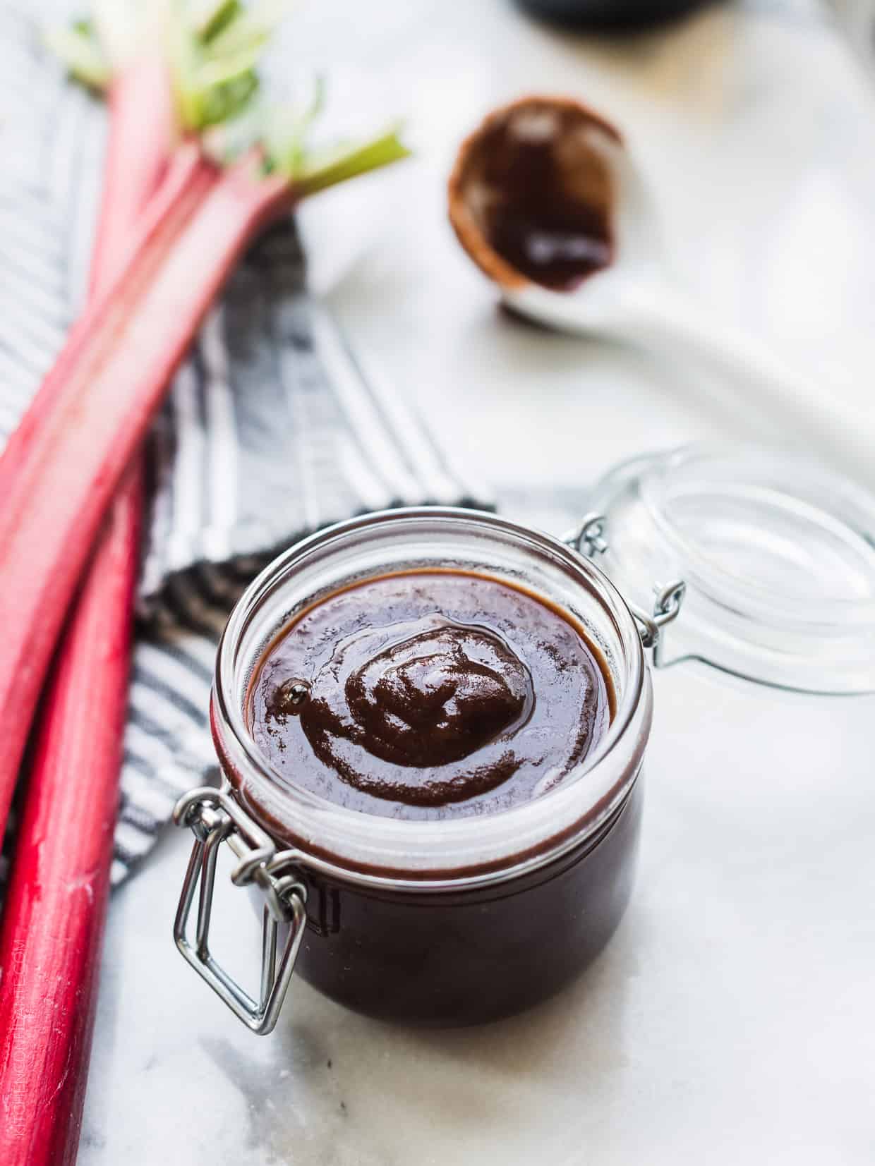 Glass jar of Rhubarb-Wine BBQ sauce surrounded by fresh stalks of rhubarb.