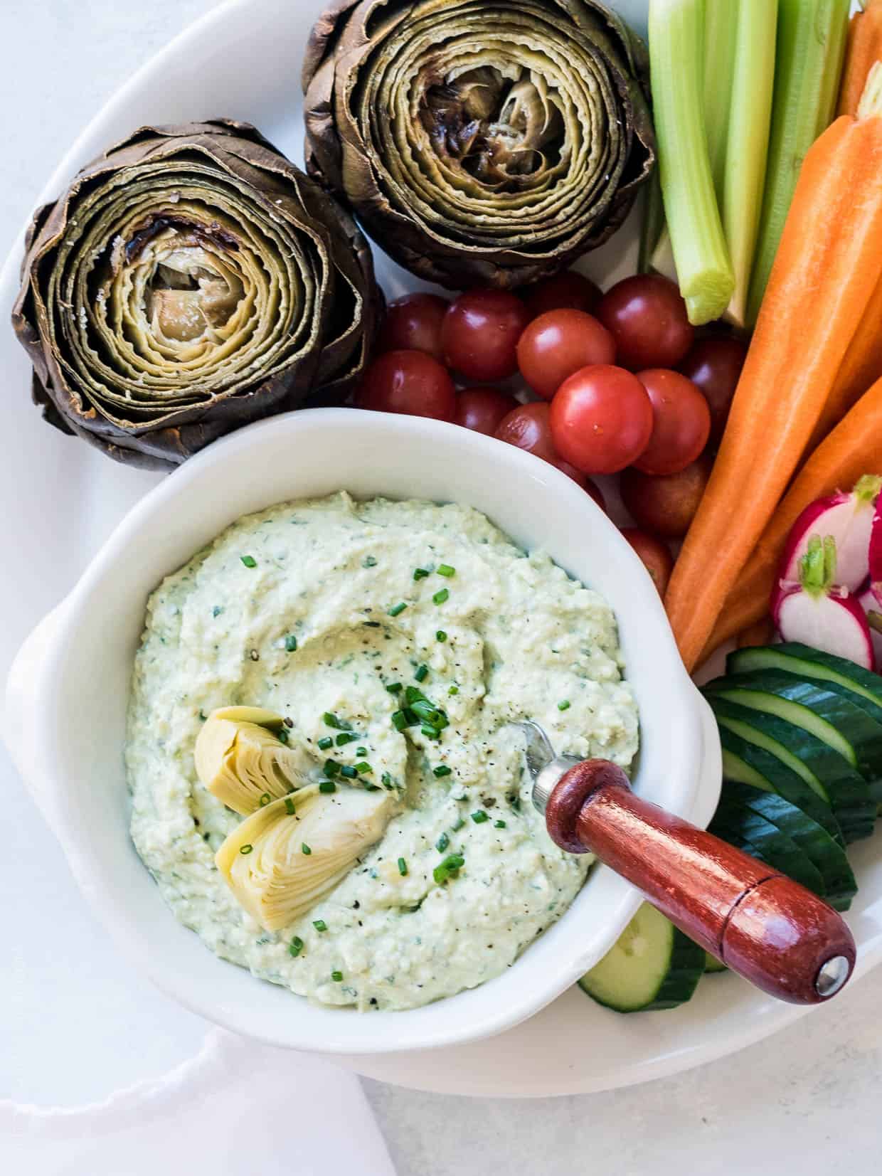 A bowl of Artichoke and Avocado Dip surrounded by vegetables including carrots, radishes, and fresh artichokes.