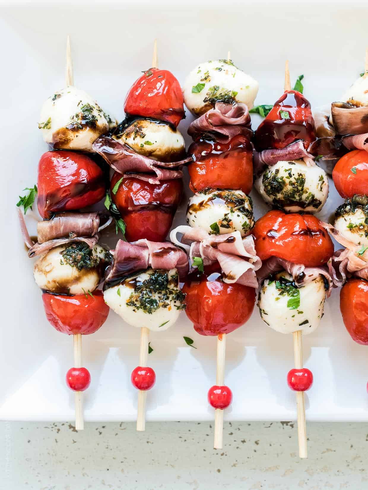 Peppadew Pepper Caprese Skewers drizzled with balsamic glaze on a white serving plate. 