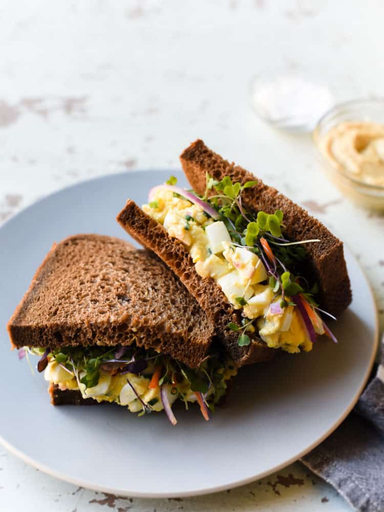 Hummus Egg Salad made into a sandwich with a layer of fresh sprouts, carrots, and red onion. The sandwich is cut in half and placed on a plate.