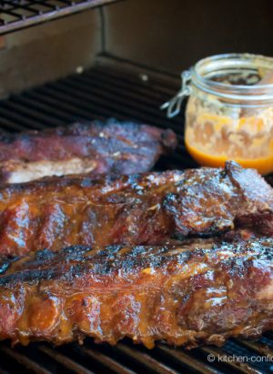 Three baby back ribs on a grill with a jar of fiery peach bbq sauce.