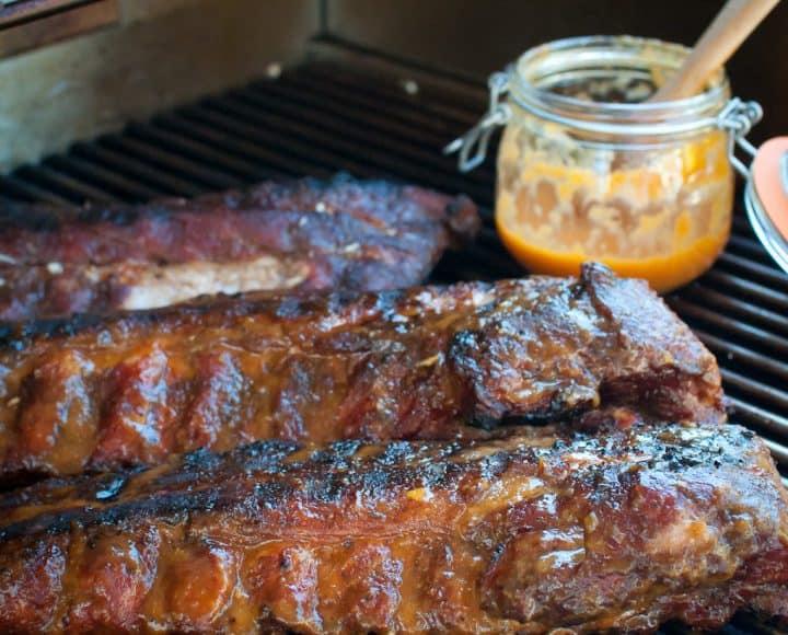 Three baby back ribs on a grill with a jar of fiery peach bbq sauce.