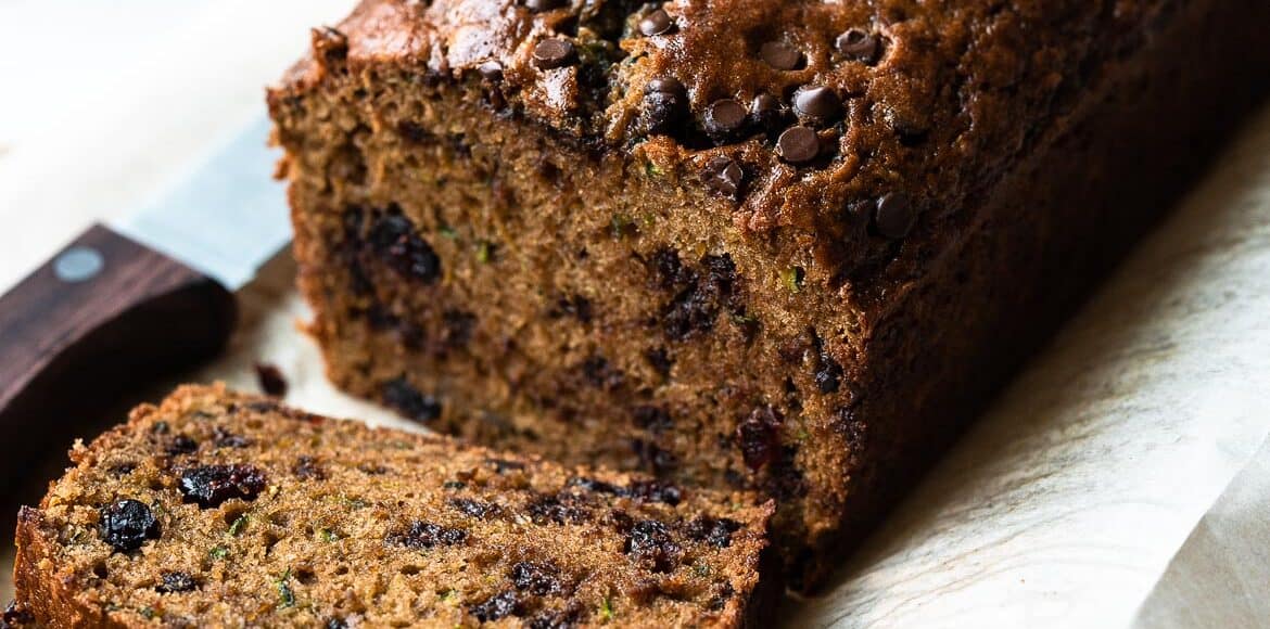 Zucchini bread with cranberries and chocolate chips sliced on a cutting board with a cup of tea in the background.