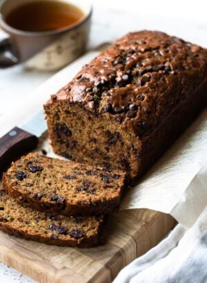 Zucchini bread with cranberries and chocolate chips sliced on a cutting board with a cup of tea in the background.