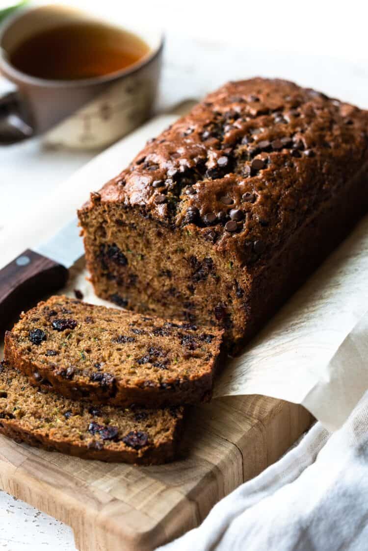 Zucchini bread with cranberries and chocolate chips sliced on a cutting board with a cup of tea in the background.