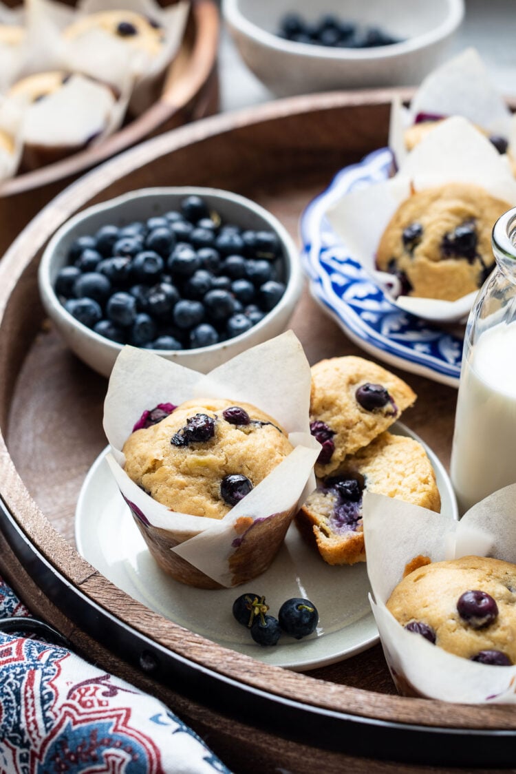 Blueberry banana muffins on a serving tray with fresh blueberries and a glass of milk.