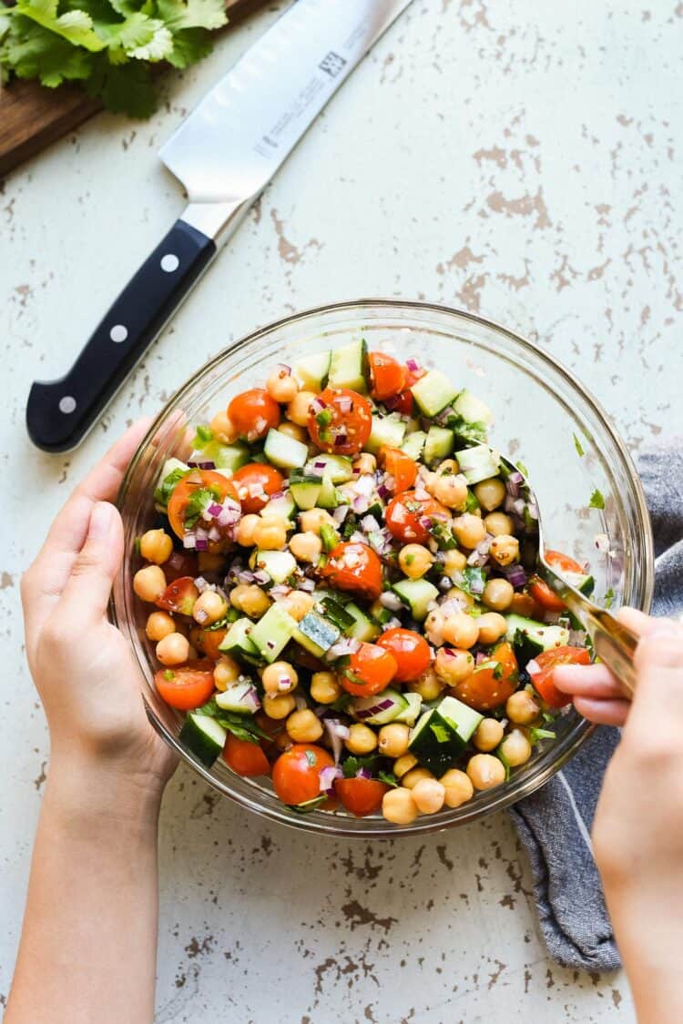 chick pea salad with tomatoes and cucumbers in a glass bowl on a rustic white background with hands holding the bowl