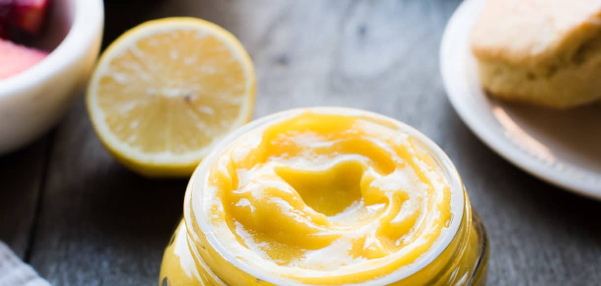 Homemade lemon curd in a glass jar on a wood table with fresh lemons and scones in the background.