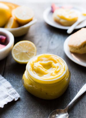 Homemade lemon curd in a glass jar on a wood table with fresh lemons and scones in the background.