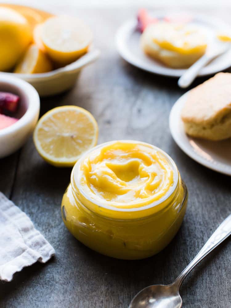 Homemade lemon curd in a glass jar on a wood table with fresh lemons and scones in the background.