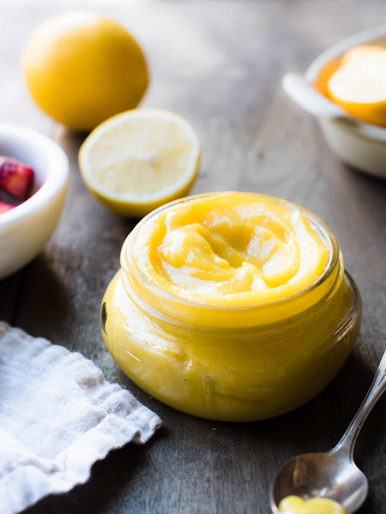 Jar of lemon curd on brown table with fresh lemon in background.