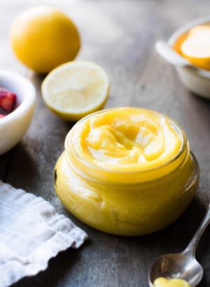Jar of lemon curd on brown table with fresh lemon in background.