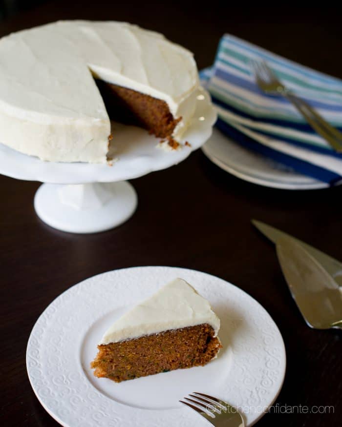 A sweet potato cake with cream cheese frosting sits on a cake stand. Beside it is a freshly cut slice sitting on a plate.