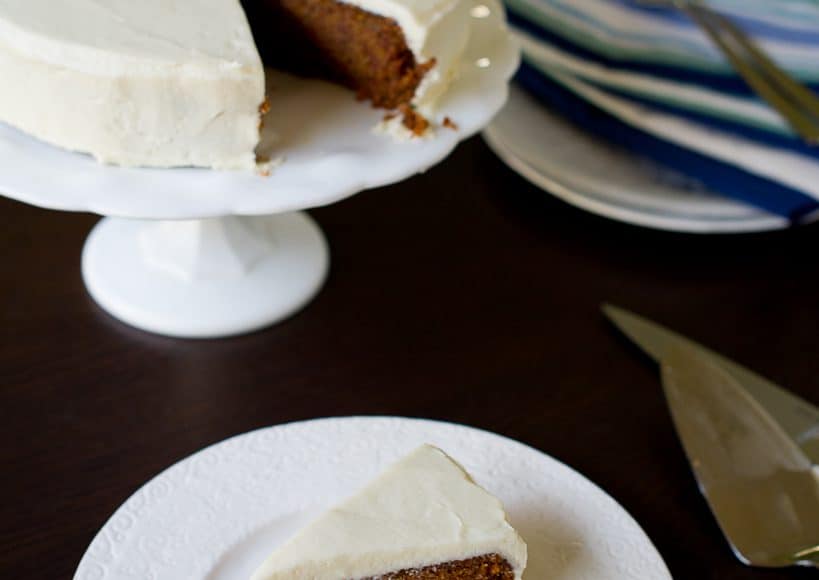 A sweet potato cake with cream cheese frosting sits on a cake stand. Beside it is a freshly cut slice sitting on a plate.