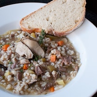 A large shallow white bowl full of beef, mushroom, and barley soup. A piece of sliced french bread sits on the edge of the bowl.