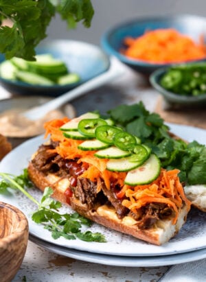 Slow Cooker Bánh Mì Vietnamese Sandwich made with slow cooker pork on a French baguette, topped with pickled carrots, cucumber slices, jalapeno and cilantro on a white plate.