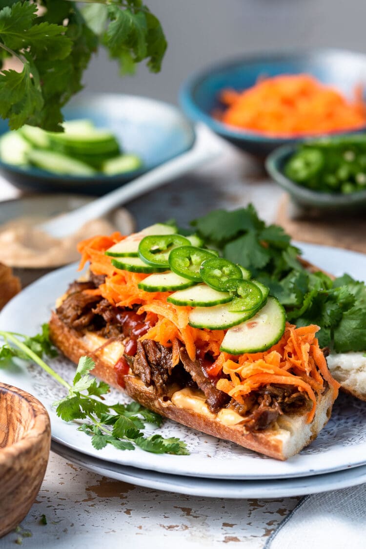 Slow Cooker Bánh Mì Vietnamese Sandwich made with slow cooker pork on a French baguette, topped with pickled carrots, cucumber slices, jalapeno and cilantro on a white plate.