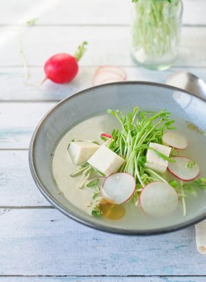 A bowl of fresh tofu soup with pea shoots and radishes floating on top.
