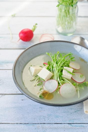 A bowl of fresh tofu soup with pea shoots and radishes floating on top.