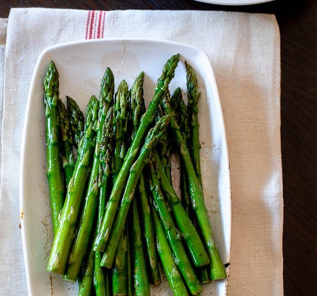 Plate of cooked asparagus on a white tile with red stripe. A bowl of pork chops is in the background.