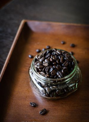 An overflowing jar of dark roasted coffee beans sits on a wooden tray.