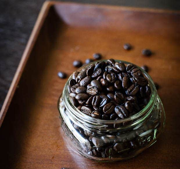 An overflowing jar of dark roasted coffee beans sits on a wooden tray.
