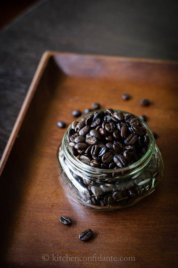 An overflowing jar of dark roasted coffee beans sits on a wooden tray.