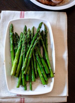Plate of cooked asparagus on a white tile with red stripe. A bowl of pork chops is in the background.