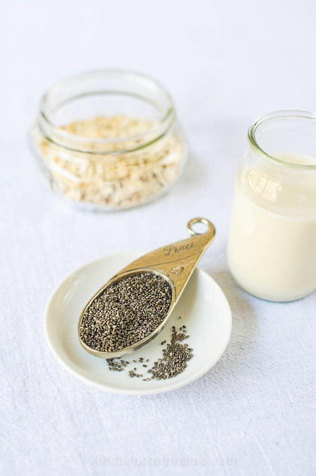 Chia seeds on a metal measuring spoon, with milk and oats in a glass mason jar in the background - ingredients for overnight refrigerator oatmeal