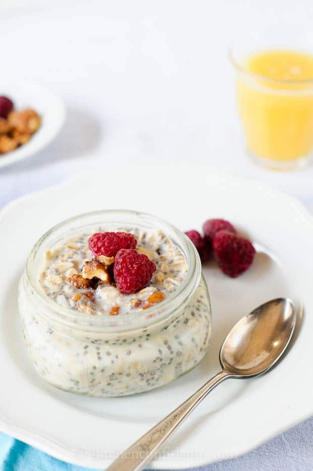 Small mason jar of overnight refrigerator oatmeal with chia seeds, walnuts, and raspberries, set on a white plate with orange juice in the background