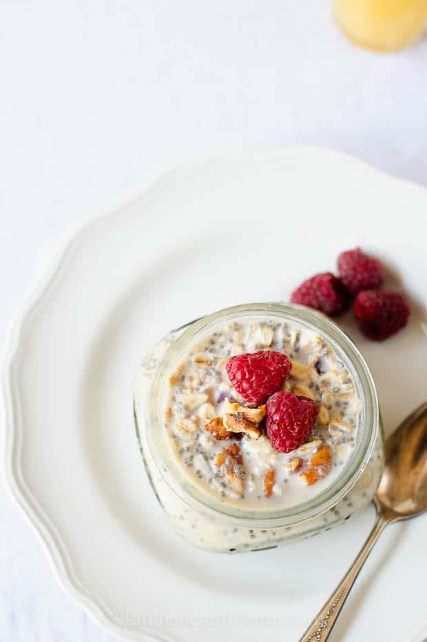 Overhead view of a small mason jar of overnight refrigerator oatmeal with chia seeds, walnuts, and raspberries, set on a white plate