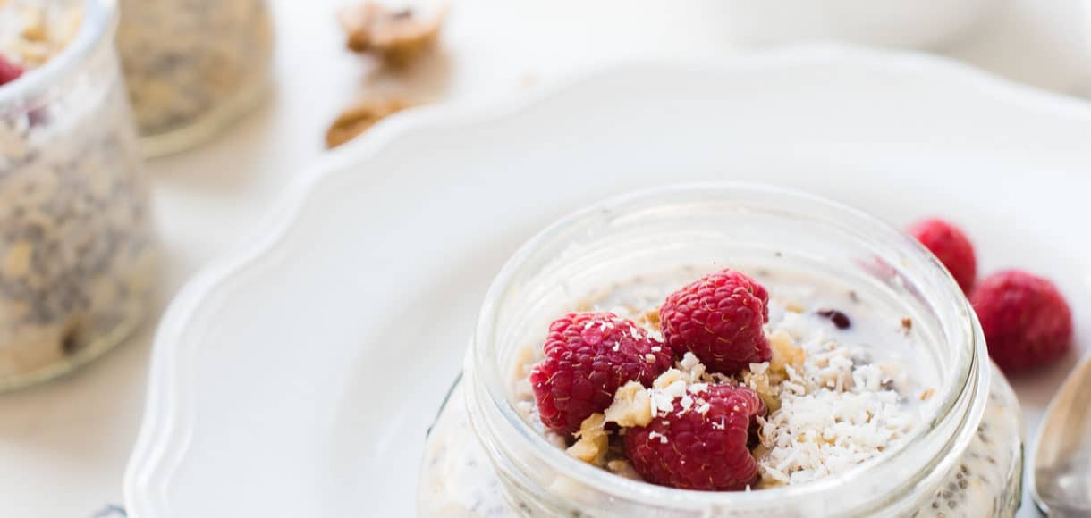 Glass mason jar of overnight refrigerator oatmeal with chia seeds, walnuts, and raspberries set on a white plate