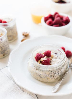Glass mason jar of overnight refrigerator oatmeal with chia seeds, walnuts, and raspberries set on a white plate