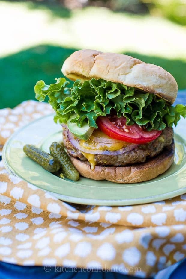 A Southwestern Turkey Burger sits on a green plate. The plate rests on a yellow and white pinic blanket lying on the grass.