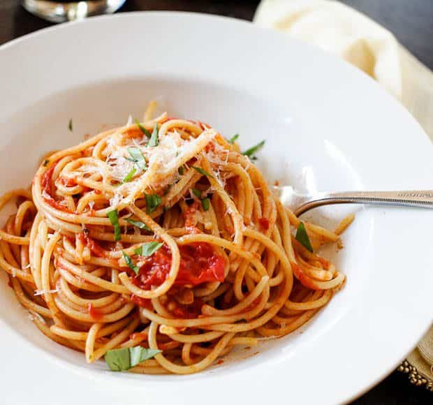 A white bowl filled with spaghetti made with homemade spaghetti sauce topped with fresh basil and parmesean reggiano cheese.