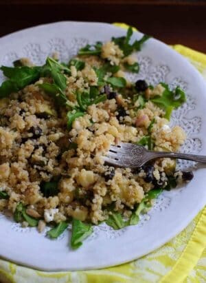 A plate of quinoa black bean salad sits on a yellow cloth. A fork lies on the plate, and corn, shallots, and arugala are mixed into the salad.