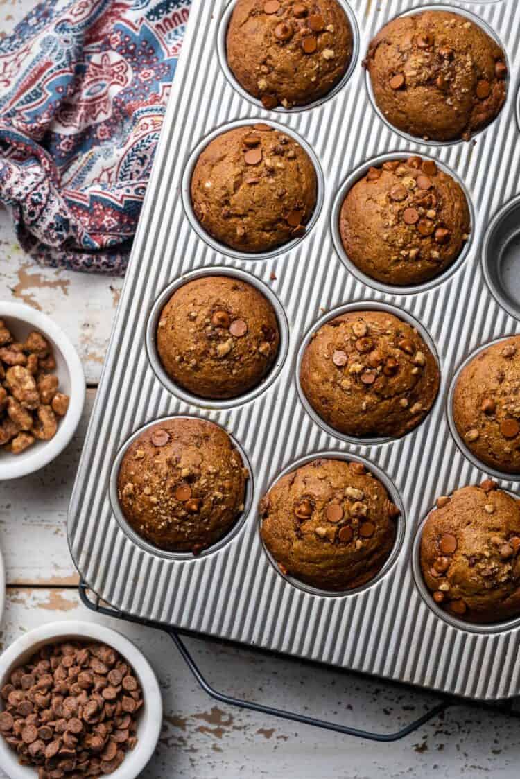 Pumpkin Cinnamon Chip & Walnut Muffins in a muffin pan with cinnamon chips and walnuts in white bowls.
