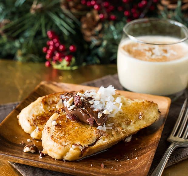 Coconut Hazelnut Eggnog French Toast sitting on a wooden plate with a glass of eggnog. In the background is wintery greenery and red berries.