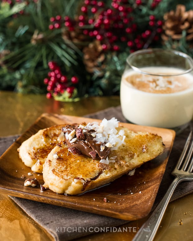 Coconut Hazelnut Eggnog French Toast sitting on a wooden plate with a glass of eggnog. In the background is wintery greenery and red berries.