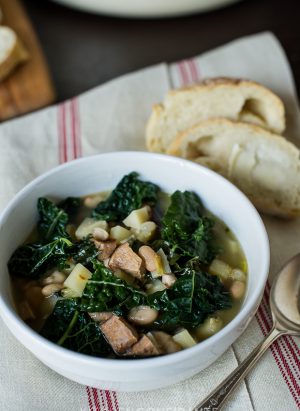 A white bowl of Kale Parsnip & Sausage Soup. The bowl sits on a white towel with red stripes. A spoon and two pieces of bread lay beside the bowl.