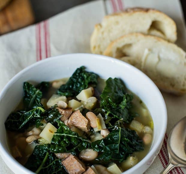 A white bowl of Kale Parsnip & Sausage Soup. The bowl sits on a white towel with red stripes. A spoon and two pieces of bread lay beside the bowl.