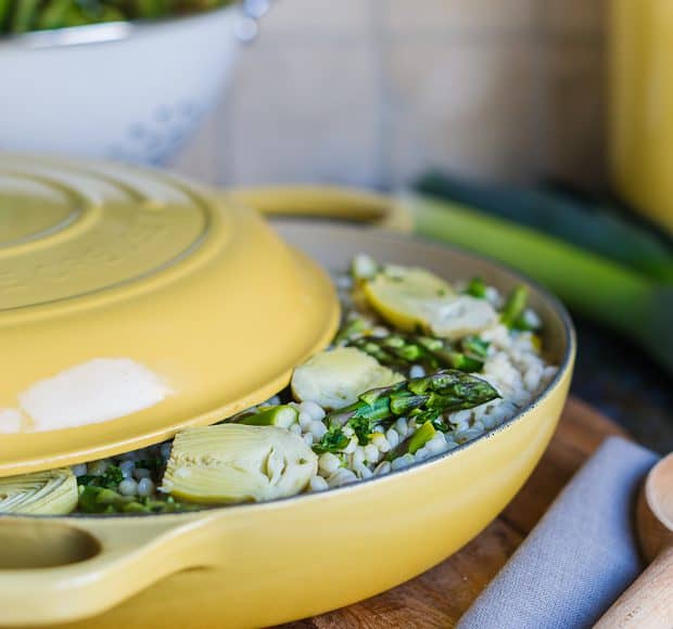 A yellow Le Creuset braising pan with lid semi-removed revealing Barley Risotto with Artichokes and Asparagus.