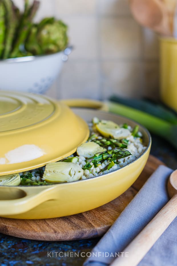 A yellow Le Creuset braising pan with lid semi-removed revealing Barley Risotto with Artichokes and Asparagus.